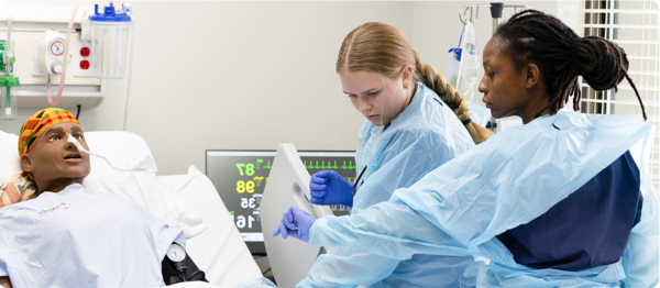 A nursing student practices their skills on a practice dummy while an instructor watches.